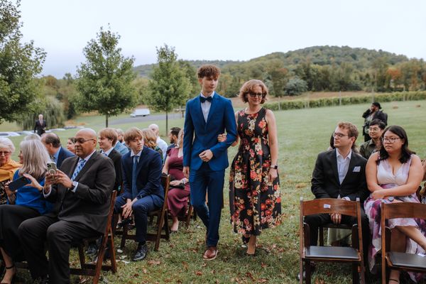 A young man in a blue suit escorts a woman in a floral dress toward the front.