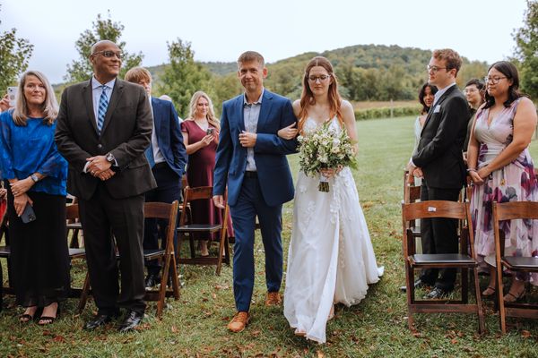On the arm of a young man in a blue suit, Anastasia walks between smiling guests.