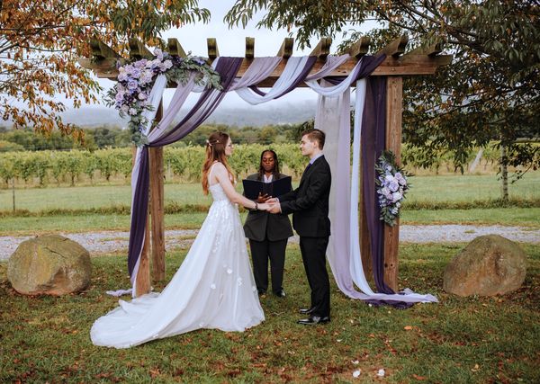 The couple faces the officiant under swaths of purple and white, the hills open behind them.