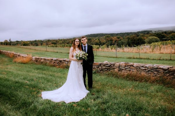 The newlyweds alone in the vineyard, a stone wall and rolling hills behind them.
